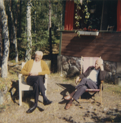Två män sitter i trädgårdsstolar utanför en stuga