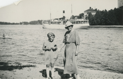 En kvinna och en flicka vid en sjö med en ångbåt i bakgrunden